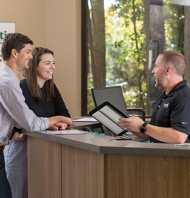 Photo of a smiling couple interacting with a friendly automotive service advisor at the front desk. The advisor is holding a car air filter while engaging with the couple in a clean, bright office with large windows showing trees outside.