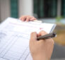 A close-up of a person filling out a car inspection checklist on a clipboard, with a blurred outdoor background.